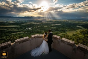 A wedding photo shows a couple on Cherokee Castle in Sedalia, Colorado. They stand atop the castle, gazing at sunlight breaking through clouds.